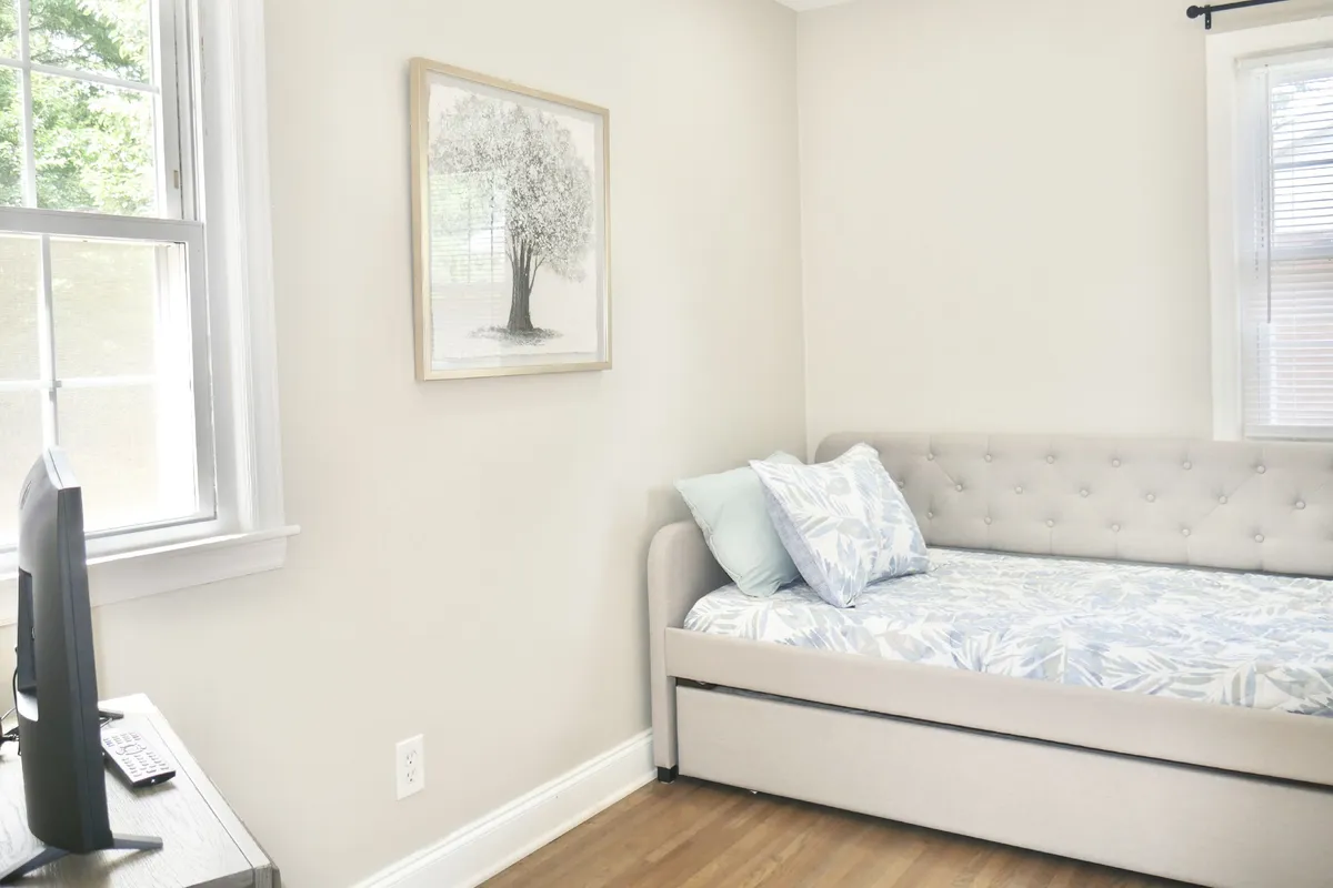 A versatile interior room featuring a daybed, wall-mounted tree artwork, and a desk setup with a monitor, ideal for both work and relaxation. The mood is calm, and the space appears adaptable to different uses.