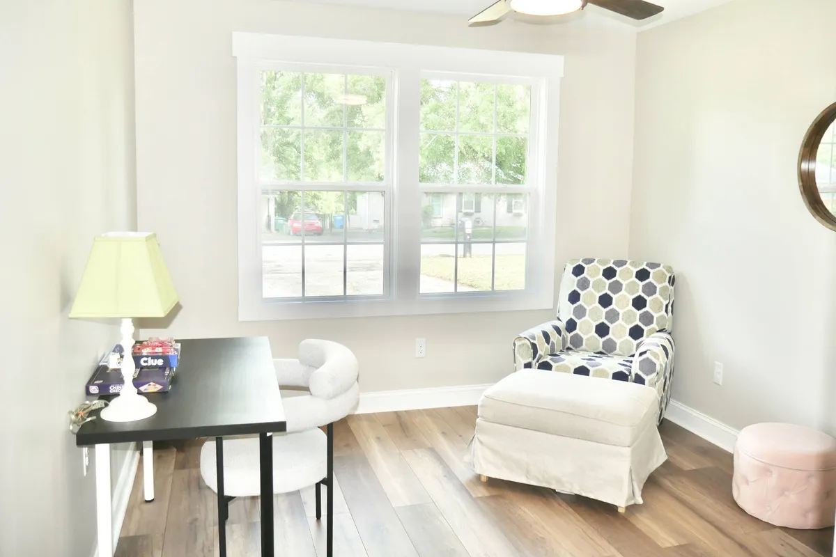 A cozy workspace featuring a black desk positioned by large windows, accompanied by a blue and white patterned chair. The room is filled with natural light, providing a serene and productive environment ideal for remote work or creative projects.