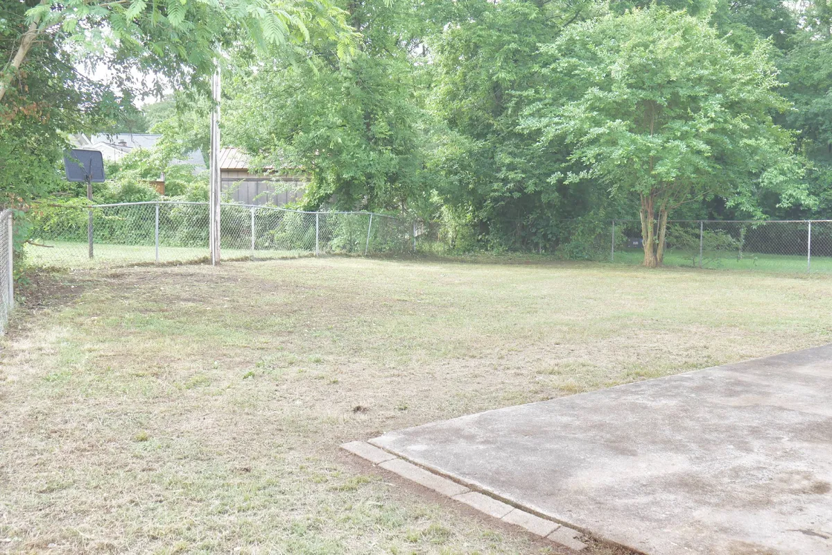 A sunlit backyard featuring a large open grassy area, bordered by a secure fence and a tree line for privacy. There's a basketball hoop for play, and ample space for various activities such as picnics, gardening, or letting pets run free. The scene conveys room for children and pets to play safely and adults to relax or entertain.