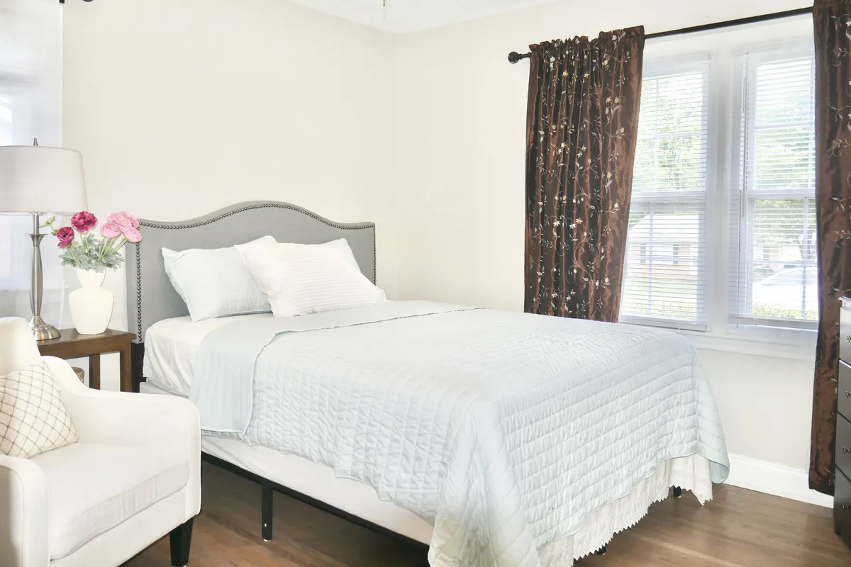 A serene bedroom featuring a comfortable bed, a cozy armchair, a lamp, and large curtained windows that let in natural light. The atmosphere is calm and inviting, ideal for relaxation or reading.