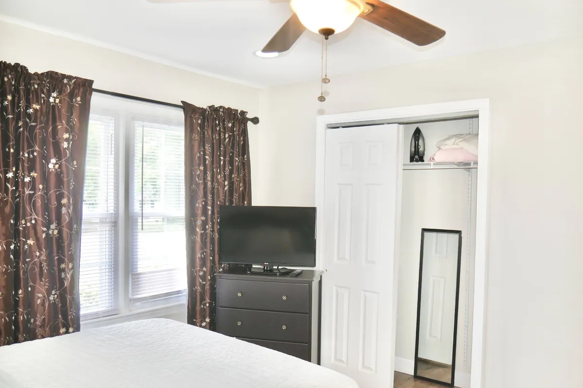 A tranquil bedroom with a neatly made bed, television, curtains, and a visible closet. The room is set up for comfort, with areas for both relaxation and entertainment.