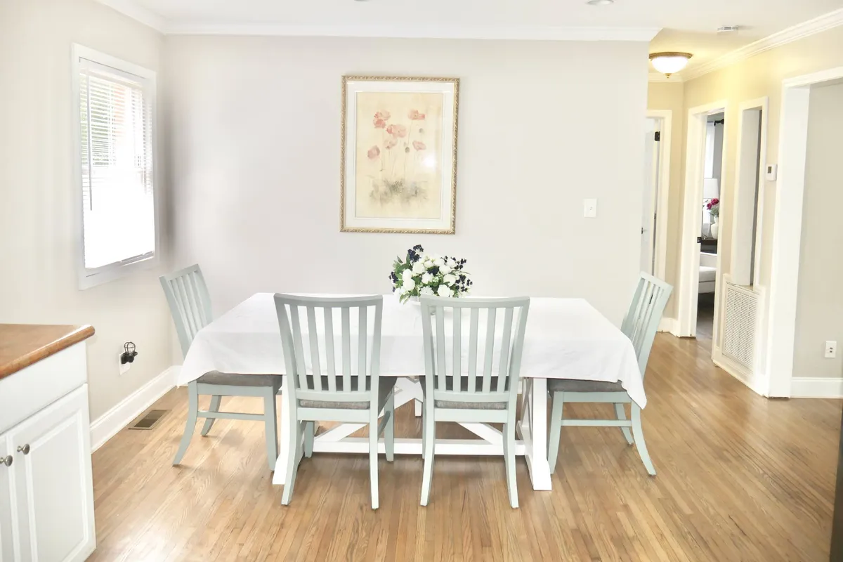 A dining area featuring a nicely set dining table adorned with a floral centerpiece and framed artwork on the wall. The setting is bright and welcoming, ideal for group meals and social gatherings.