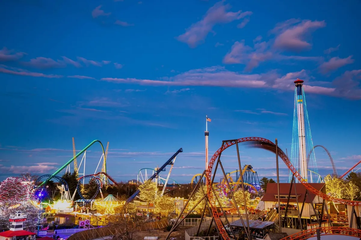A lively amusement park scene with roller coasters twisting in the background, bright carnival lights illuminating the area, crowds enjoying rides, and food stalls offering treats. The energy is palpable with families and friends taking photos and laughing together.